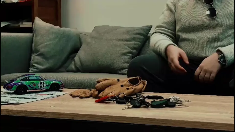 Un passionné assis sur un canapé avec des gants de conduite et une miniature de Porsche 911